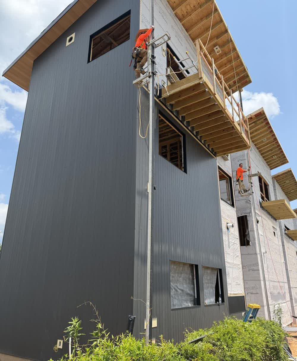 A Circuit Villa clad in dark gunmetal siding, cantilevered deck framing above, with crews working from lifts alongside
