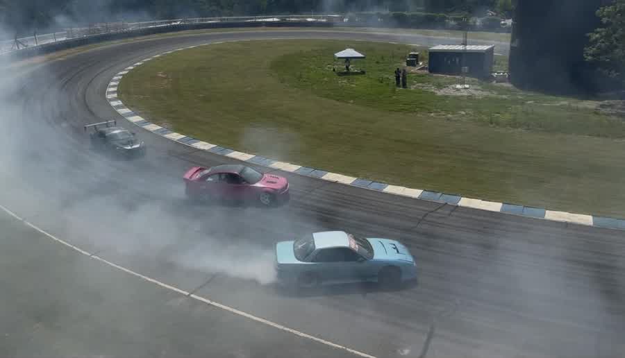 Three cars drifting in formation through the Carousel, the corner filled with tire smoke, seen from balcony level