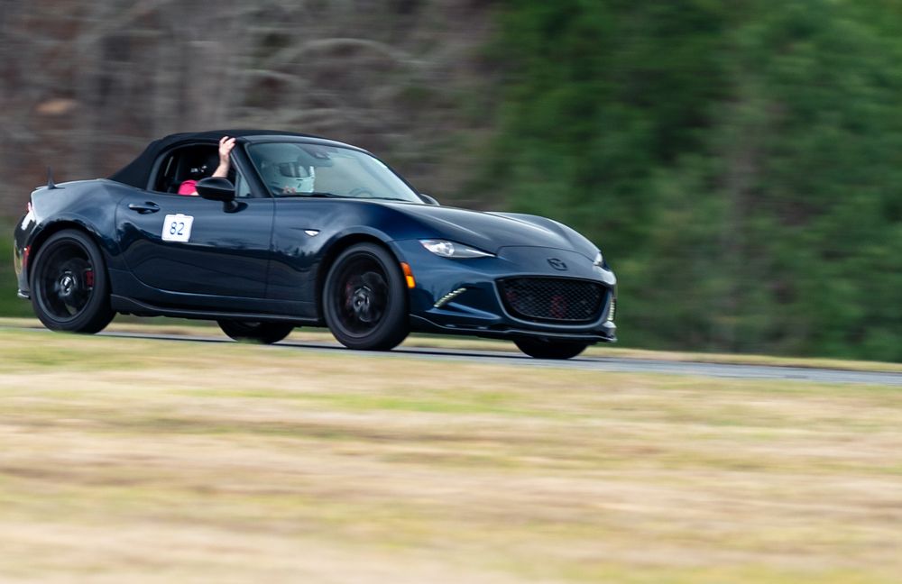 A two-seat sports car on track with two helmeted occupants, the instructor's arm raised