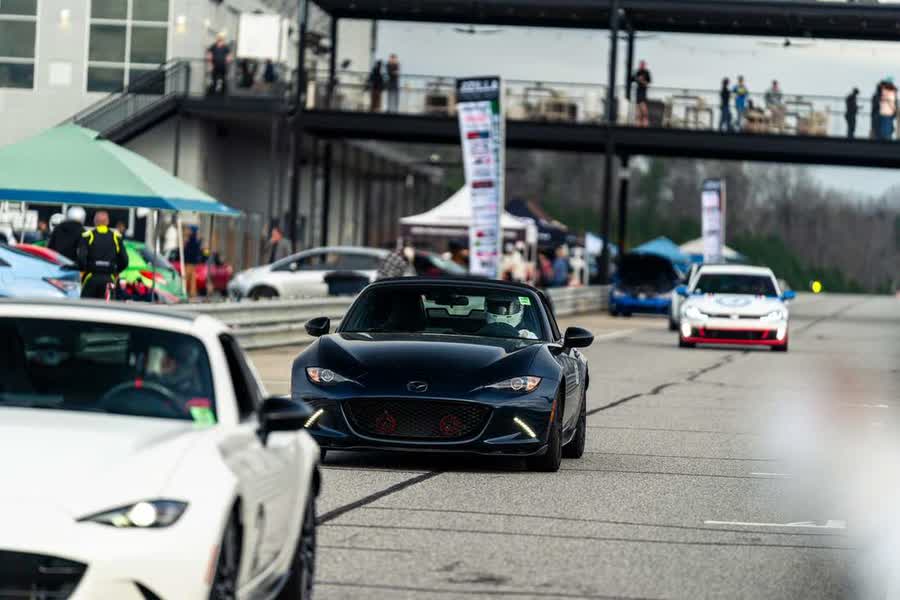 Cars queued on pit lane at an HPDE day, with paddock tents, competitors and the clubhouse behind