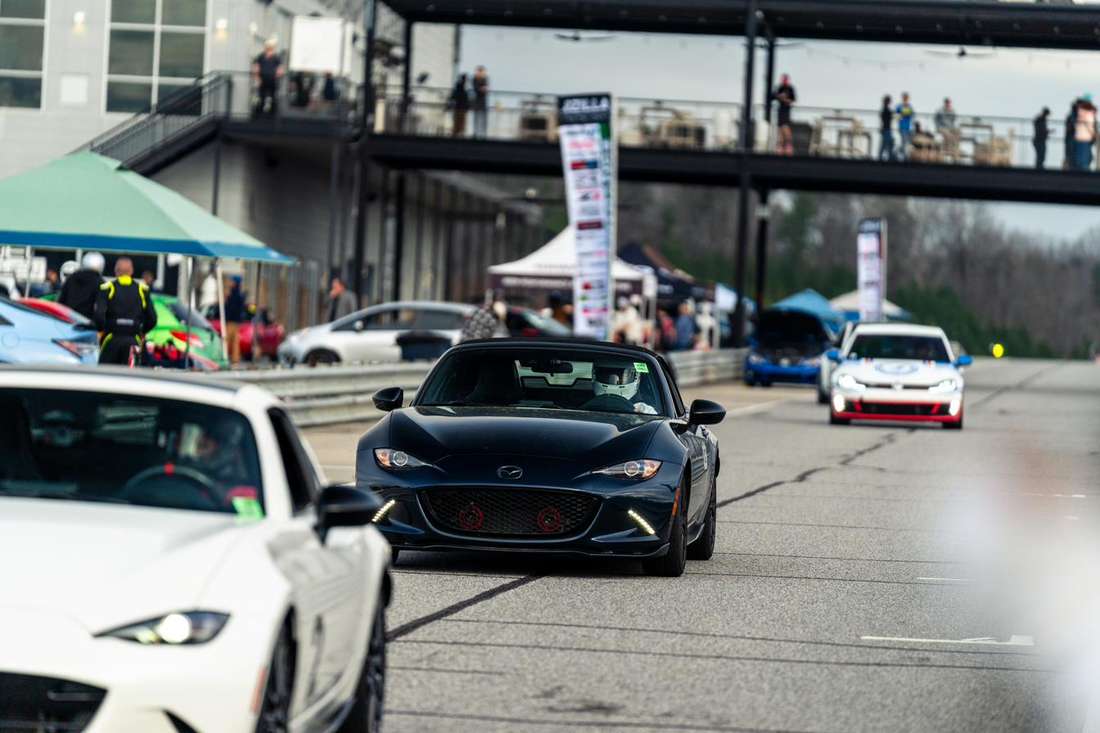 Cars queued on pit lane at an HPDE day, with paddock tents, competitors and the clubhouse balcony behind